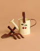 The Konges Sloejd Garden Set, featuring a cream-colored watering can with cherry design and brown handle, sits with a matching brown hand spade and fork, making a charming kids' gardening set on a light brown background.
