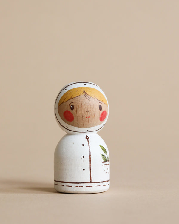 A handmade wooden astronaut painted to look like a girl with blonde hair and rosy cheeks, wearing a white shirt with a green leaf pattern, set against a plain beige background, measures 2.2