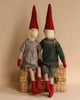 Two Maileg Christmas Pixy (Size 5) dolls with pointed red hats sitting side by side on a wicker basket, against a neutral background. The dolls feature striped leggings, intricate clothing made from exclusive materials, and stitched