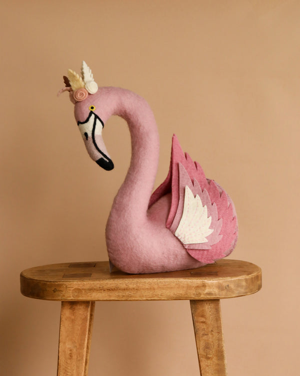 The Handmade Felt Flamingo with Wings, crafted in Nepal from soft pink felt and detailed with white wings and a crown, sits on a wooden stool against a plain beige background.
