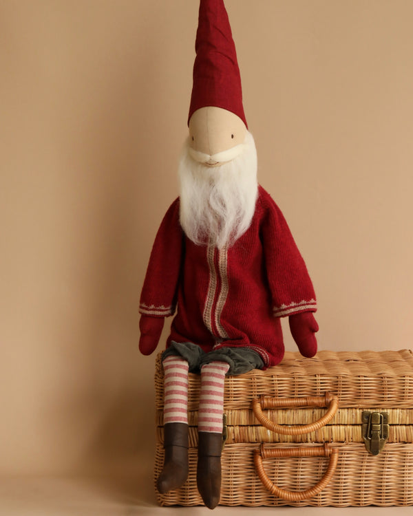 A Maileg Small Santa doll sits on a wicker basket, wearing a red lambswool sweater with white detailing, striped pants, and brown shoes, against a neutral beige background.
