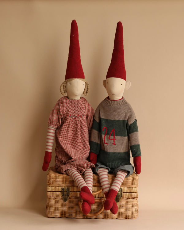 Two Maileg Christmas Pixy (Size 6) dolls with red hats are sitting on a wicker basket. One wears a red checkered outfit, and the other wears a grey sweater with the number 24. Both are designed as
