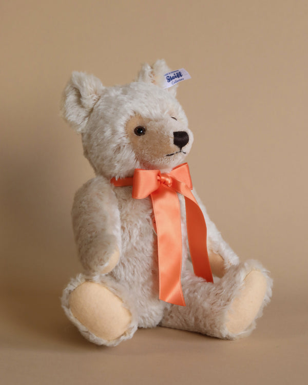 A fluffy, beige Classic Mohair Teddy Bear with a bright orange ribbon tied around its neck sits against a tan background, featuring a blue "Steiff" tag on its ear.