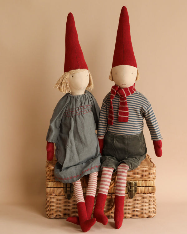 Two Maileg Christmas Small Pixy dolls with elongated red hats are seated on a wicker basket. One wears a grey dress; the other, a blue and red striped shirt with striped leggings.