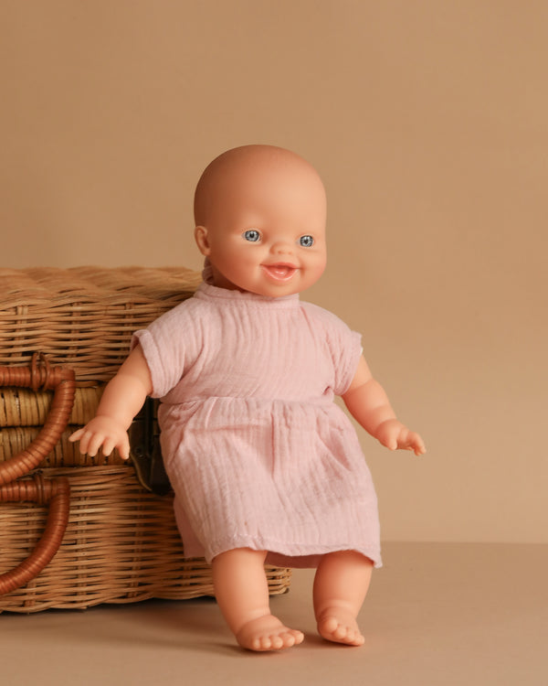 The Minikane Soft Body Doll (11") - Liv with Clothing, featuring blue eyes and a light pink dress, sits upright next to a wicker basket against a beige background.