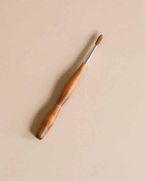A Stockmar Paint Brush - Round Tip with a wooden handle, silver ferrule, and round, pointed brush tip lies centered on a plain beige background.