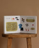 Our Big Book of Sports: A Collection of 100+ Sports Words sits open on a stool, showing vivid pictures of soccer gear like field, jersey, ball, gloves, and shin guards—perfect for teaching sports vocabulary to preschoolers against a beige backdrop.