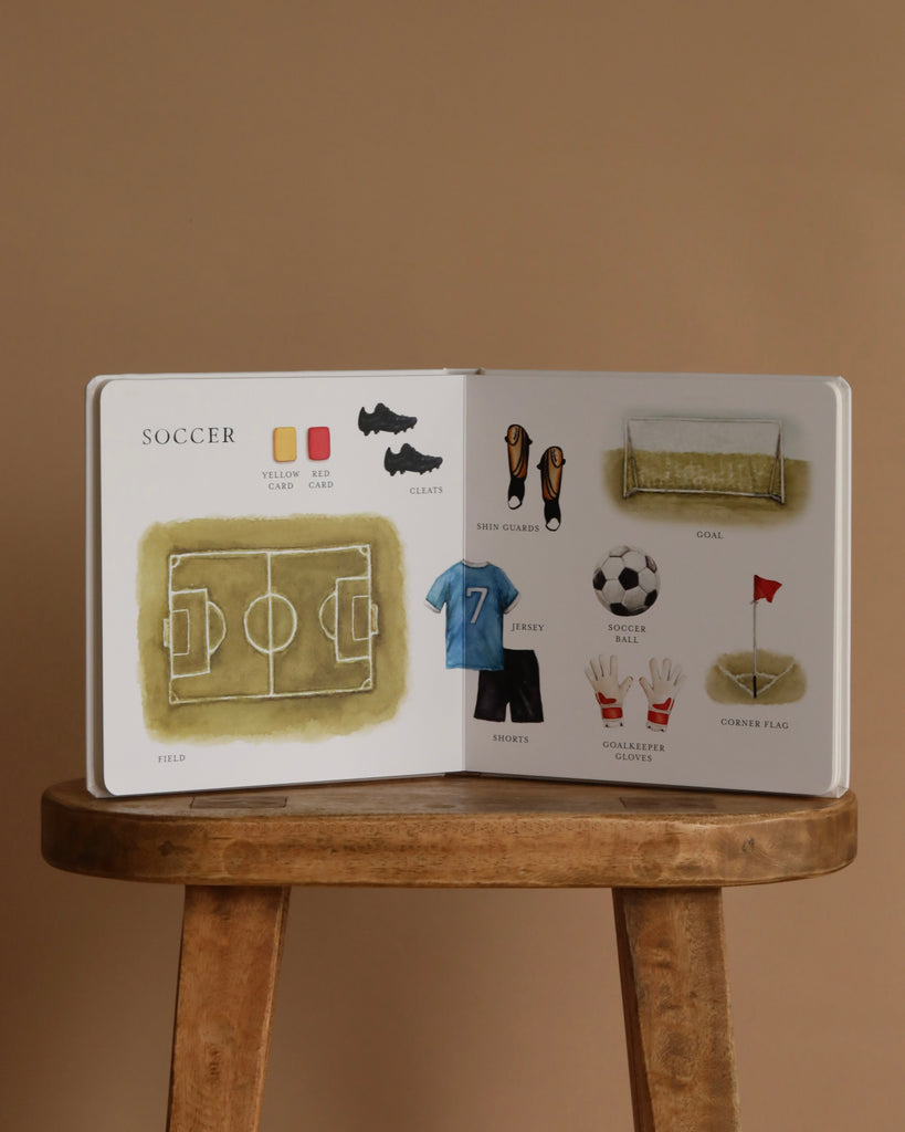 Our Big Book of Sports: A Collection of 100+ Sports Words sits open on a stool, showing vivid pictures of soccer gear like field, jersey, ball, gloves, and shin guards—perfect for teaching sports vocabulary to preschoolers against a beige backdrop.