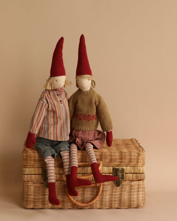 Two Maileg Christmas Pixy (Size 4) dolls with red hats, crafted from exclusive materials, seated on a wicker suitcase against a neutral background. One doll wears a striped shirt and shorts, the other a knit sweater and pants.