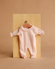 A pale pink long-sleeve onesie with ruffled collar, perfect for the Minikane Soft Body Doll (11") - Claire with Clothing, displayed on a wooden hanger against a light wood and beige background.