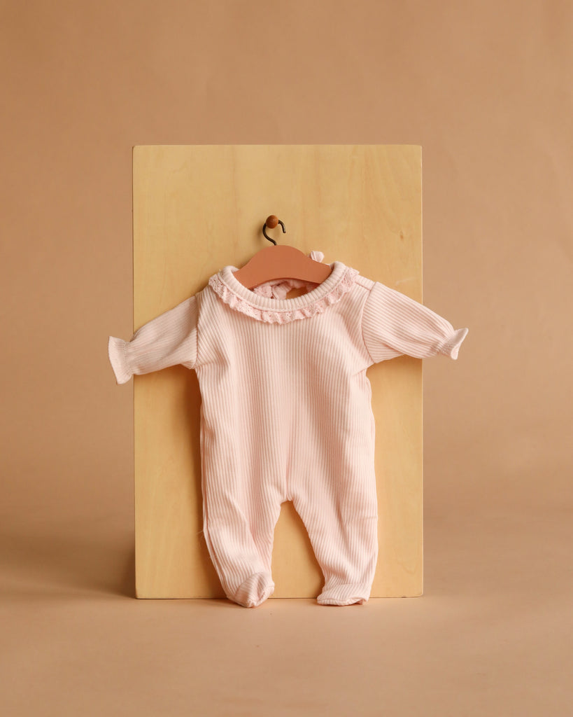 A pale pink long-sleeve onesie with ruffled collar, perfect for the Minikane Soft Body Doll (11") - Claire with Clothing, displayed on a wooden hanger against a light wood and beige background.