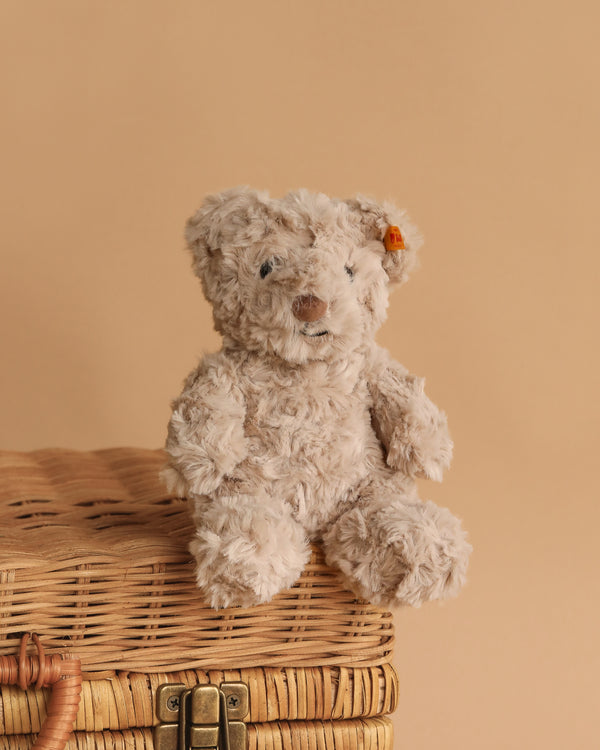 The Steiff Honey Teddy Bear Stuffed Plush, 8 Inches, with a small orange ear tag, sits on a closed wicker basket against a beige background, creating an adorable children’s toy display.