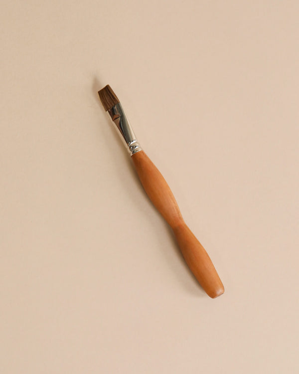 A single Stockmar Paint Brush - Flat Tip with a natural wooden handle and silver ferrule is lying on a smooth, beige background.