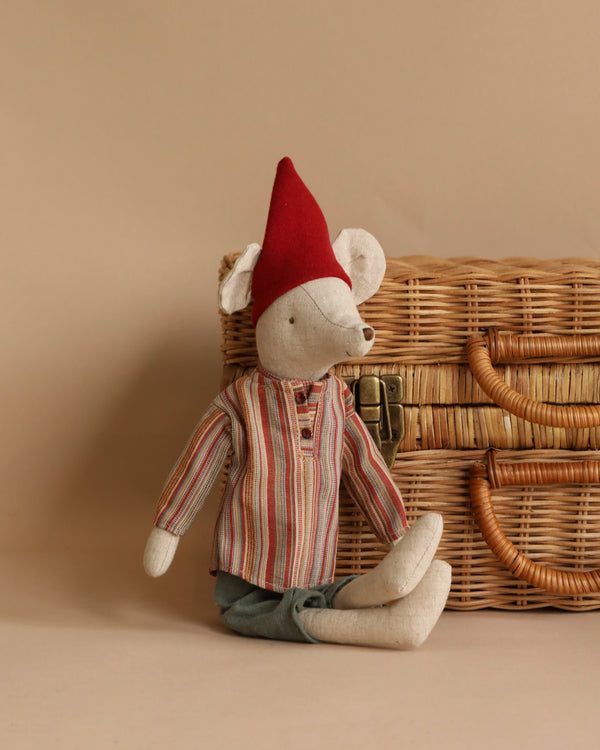 A Maileg Medium Christmas Mouse - Boy wearing a red pointed hat and cozy Christmas clothes, sitting in front of a wicker basket on a neutral background.