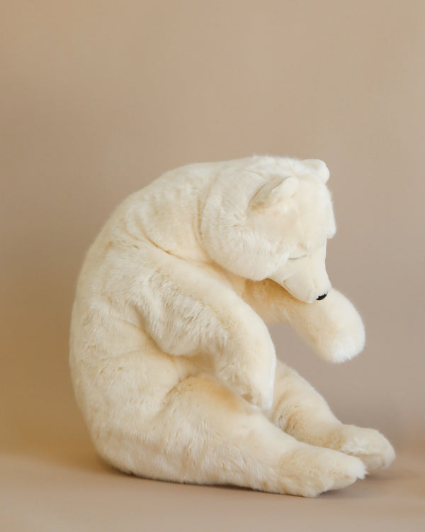 A Sleeping Polar Bear Stuffed Animal, hand sewn with artisanal craftsmanship, sitting with its head bowed and arms wrapped around its knees, displayed against a soft beige background.