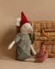 A Maileg Medium Christmas Mouse - Girl doll wearing a red hat, checkered shirt, and striped socks, sitting next to a wicker basket on a beige background.