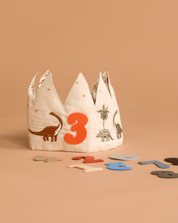 The Konges Sloejd Birthday Crown - Dino features embroidered dinosaurs and a bold orange number 3, with colorful felt numbers. It's a fun, adjustable birthday accessory, shown on a beige surface and background.