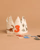 The Konges Sloejd Birthday Crown - Dino features embroidered dinosaurs and a bold orange number 3, with colorful felt numbers. It's a fun, adjustable birthday accessory, shown on a beige surface and background.