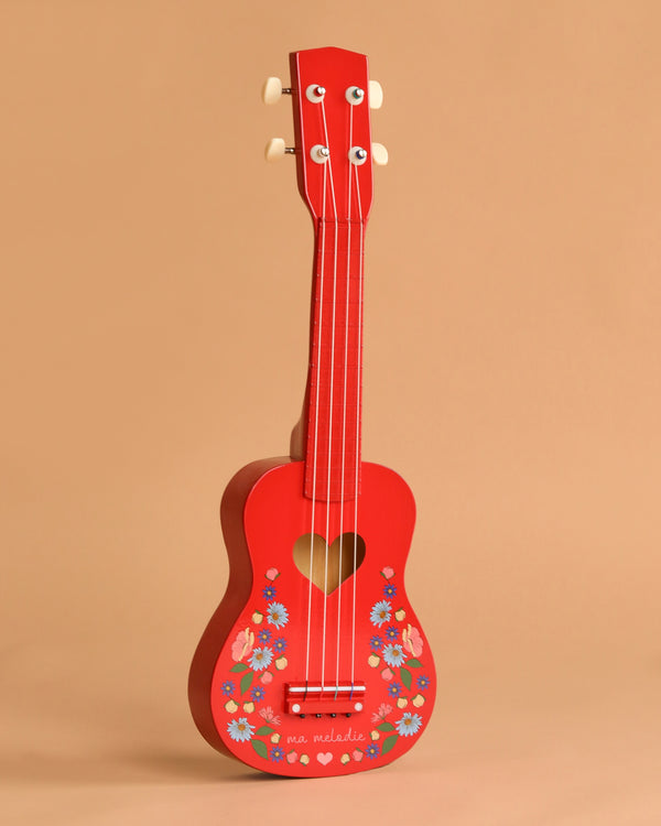 The Konges Sloejd Wooden Ukulele - Barbados Cherry, a small red ukulele for kids with a heart-shaped sound hole and floral details, stands upright on a plain beige background. “ma mélodie” is inscribed near its base.
