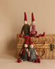 Four Maileg Christmas Mini Pixy dolls (Size 2) with red hats and shoes sit on and around a wicker suitcase against a beige background. Dressed in cozy outfits, including sweaters, overalls, and dresses made from exclusive materials, they evoke a charming, handcrafted vibe—perfect as delightful Christmas decorations.