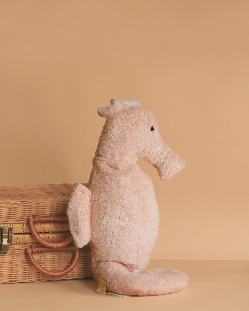 The Senger Naturwelt Cuddly Animal - Seahorse in light pink sits upright beside a closed wicker basket with handles, set against a plain beige background.