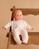 The Minikane Soft Body Doll (11") - Claire with Clothing, a phthalate-free vinyl doll with light skin and short molded hair in a white one-piece, sits on a wooden chair against a beige background.