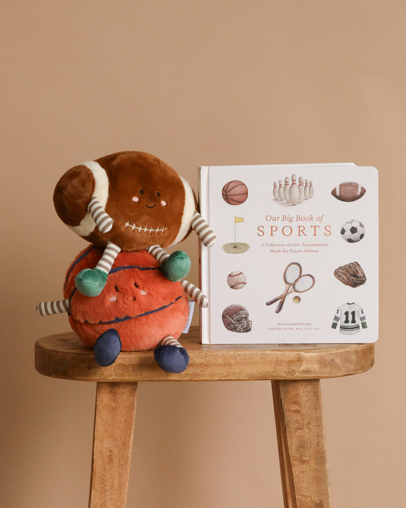 The Sports Storytime Set featuring plush football and basketball toys rests on a wooden stool beside an illustrated preschool sports book, all set against a beige background.