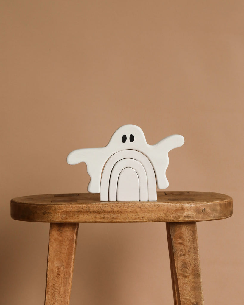 The Handmade Wooden Ghost Stacker, a whimsical white wooden toy with black eyes and layered arches for sensory play, is displayed on a wooden stool against a plain beige background.