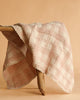 The Organic Cotton Baby Blanket - French Plaid, Petal features soft pink and white plaid with lace-trimmed edges, draped over a rustic wooden bench set against a beige background.