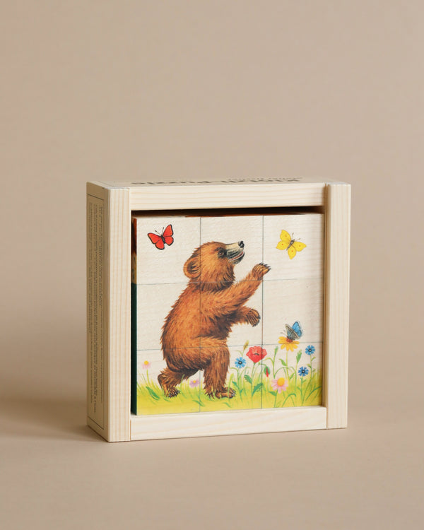 A small, Wooden Block Puzzle - 9-Piece Bear made in Switzerland depicting a bear standing on grass, reaching out to colorful butterflies, surrounded by flowers. The book's edges are visible, suggesting multiple pages.