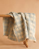 The Organic Cotton Baby Blanket - French Plaid, Blue, featuring soft blue and white plaid with lace trim and made in Portugal, is draped over a rustic wooden bench against a beige backdrop.