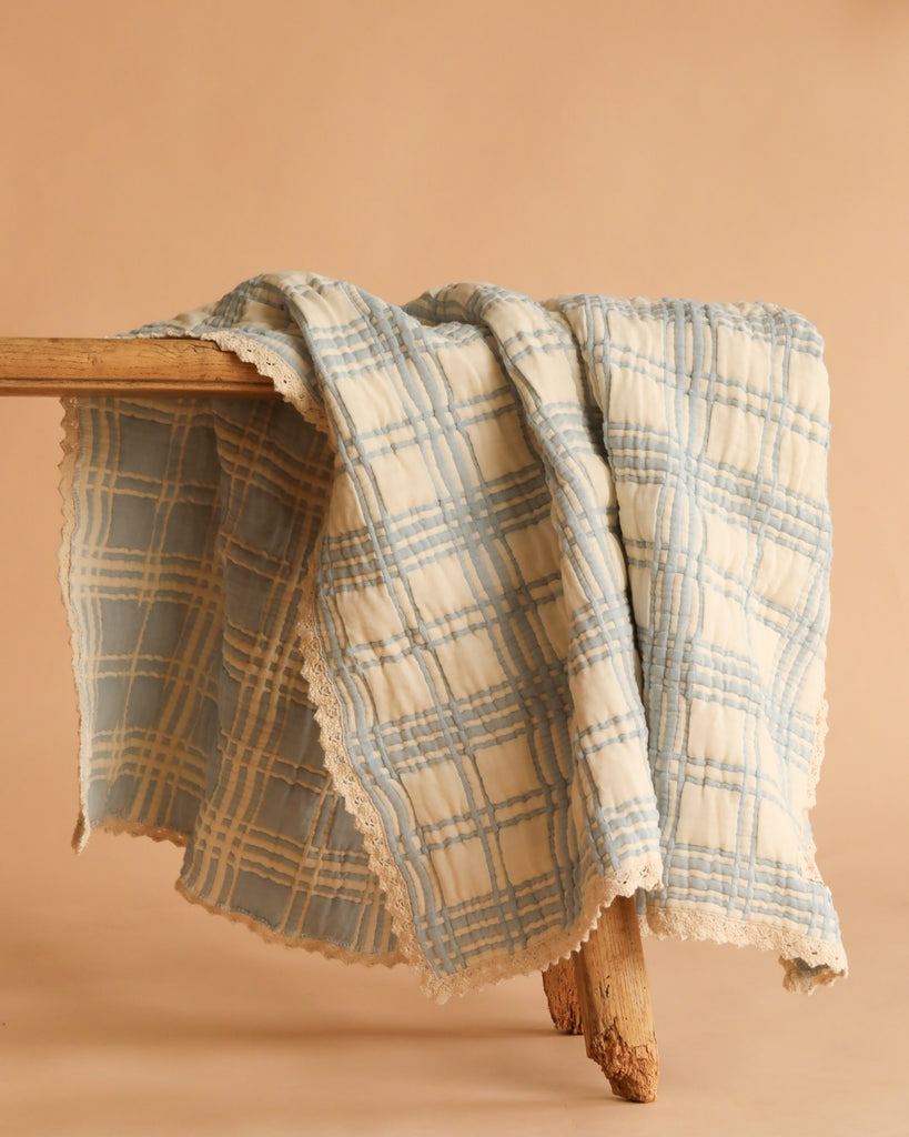 The Organic Cotton Baby Blanket - French Plaid, Blue, featuring soft blue and white plaid with lace trim and made in Portugal, is draped over a rustic wooden bench against a beige backdrop.