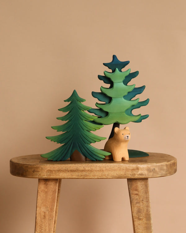 The Bumbu Handmade Wooden Bear and Forest Trees Set, crafted from sustainably sourced wood, includes two green-painted trees and a light tan bear, displayed simply on a stool against a beige background.