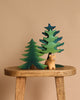 The Bumbu Handmade Wooden Bear and Forest Trees Set, crafted from sustainably sourced wood, includes two green-painted trees and a light tan bear, displayed simply on a stool against a beige background.