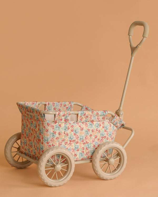 The Konges Sloejd Stoller Wagon - Vienna is a beige kids' stroller wagon with floral fabric, four large wheels, and a curved handle, shown against a plain peach background.