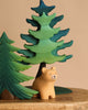 The Bumbu Handmade Wooden Bear and Forest Trees Set features a small wooden bear crafted from sustainably sourced wood, placed before two painted trees—one dark green, one light green—on a wooden surface with a beige background.