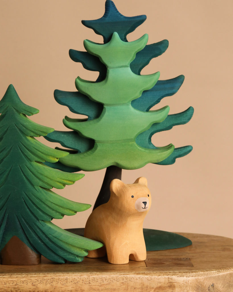 The Bumbu Handmade Wooden Bear and Forest Trees Set features a small wooden bear crafted from sustainably sourced wood, placed before two painted trees—one dark green, one light green—on a wooden surface with a beige background.
