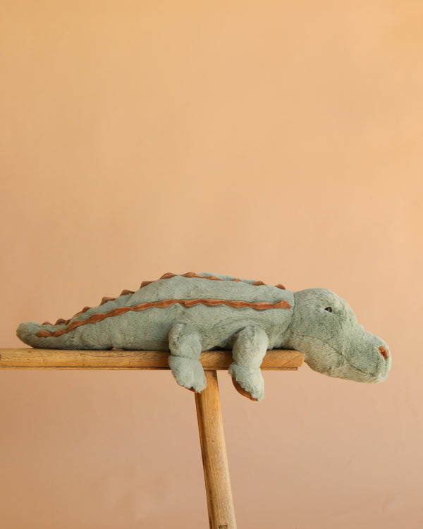 A soft, green Bukowski - The Great Maximus Alligator stuffed animal with brown accents lies stretched out on a wooden plank against a peach-colored background.