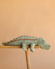 A soft, green Bukowski - The Great Maximus Alligator stuffed animal with brown accents lies stretched out on a wooden plank against a peach-colored background.