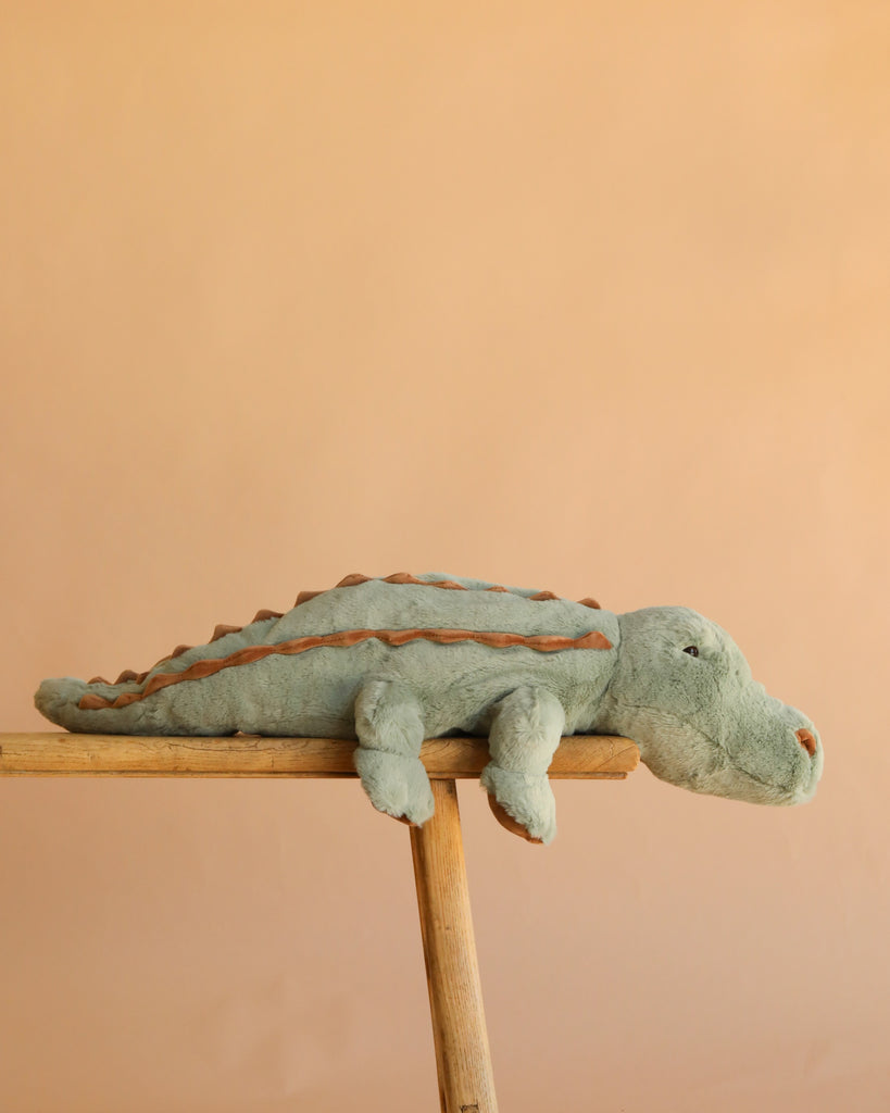 A soft, green Bukowski - The Great Maximus Alligator stuffed animal with brown accents lies stretched out on a wooden plank against a peach-colored background.