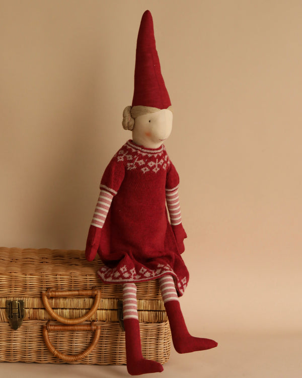A Maileg Small Mrs. Clause (Size: 33 in.) dressed as Mrs. Claus with a pointed red hat and patterned sweater dress, sitting on a wicker basket against a beige background. The doll has simplistic facial features and striped limbs.