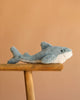 The Bukowski - Splash Shark, a soft toy in calming blue and white, sits on a wooden stool set against a simple light brown background.