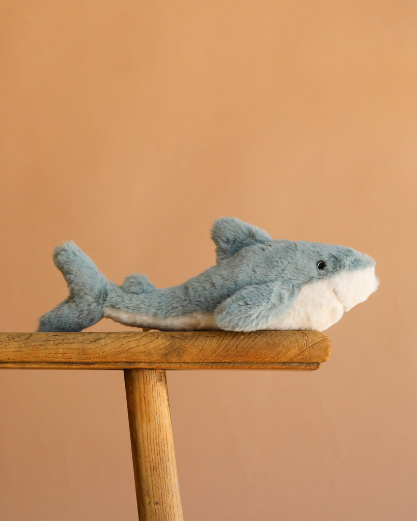 The Bukowski - Splash Shark, a soft toy in calming blue and white, sits on a wooden stool set against a simple light brown background.