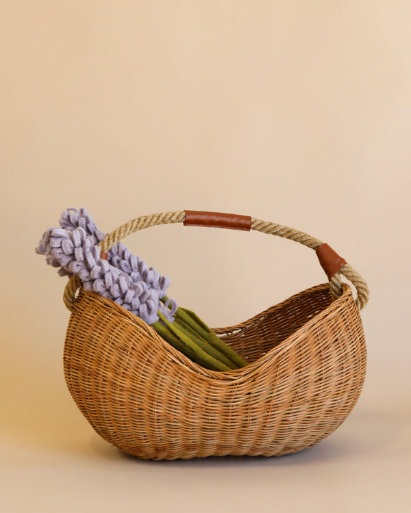 A Basket of Lavender Felt Flowers with a rope handle, partly wrapped in leather, sits on a beige background. The natural rattan basket holds a bunch of light purple flowers with green stems, elegantly arranged to one side.