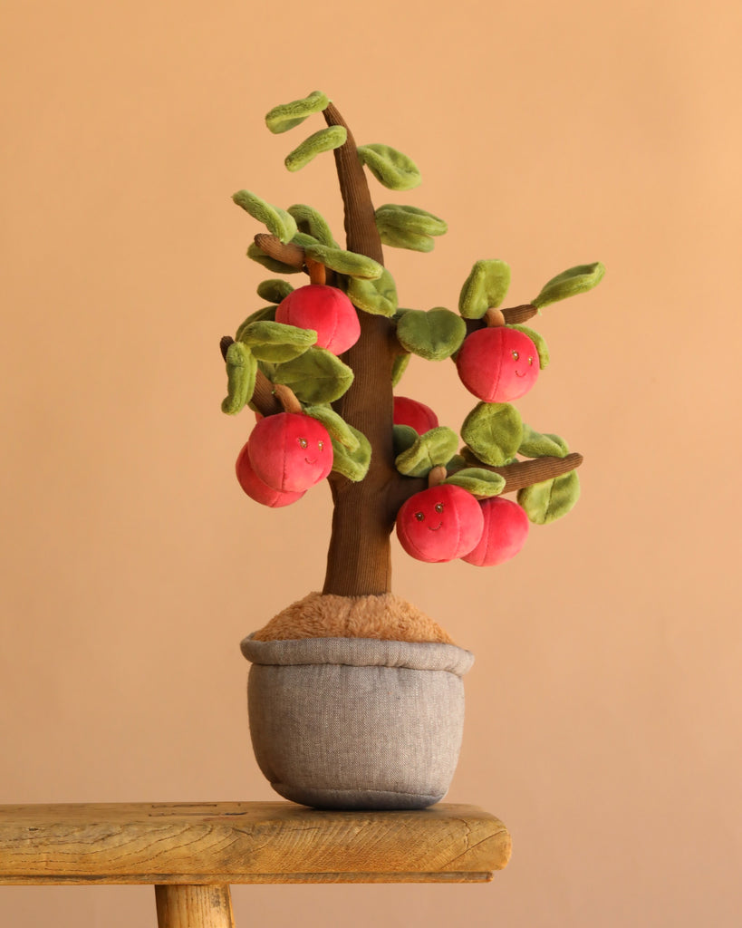 The Bukowski - Apple Tree is a plush toy featuring green leaves and red, smiling fruit faces in a fabric pot, set on wood against a plain beige background. Perfect for imaginative play and designed with safety in mind.