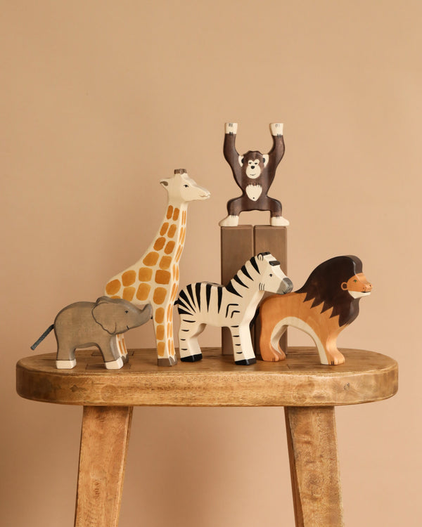 Five animals from the Holztiger Handmade Wooden Safari Animals Set—a giraffe, monkey, elephant, zebra, and lion—are arranged on a wooden stool with a beige background. The monkey stands raised on a small block for added playful charm.