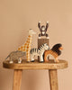 Five animals from the Holztiger Handmade Wooden Safari Animals Set—a giraffe, monkey, elephant, zebra, and lion—are arranged on a wooden stool with a beige background. The monkey stands raised on a small block for added playful charm.