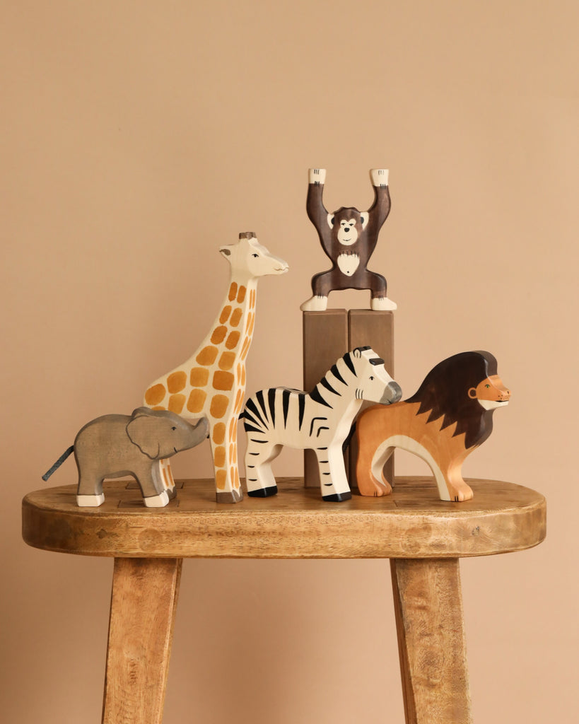 Five animals from the Holztiger Handmade Wooden Safari Animals Set—a giraffe, monkey, elephant, zebra, and lion—are arranged on a wooden stool with a beige background. The monkey stands raised on a small block for added playful charm.