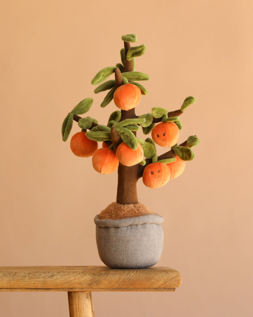 The Bukowski - Orange Tree is a plush toy featuring green leaves and smiling orange fruit in a soft gray pot, displayed on a wooden surface with a beige background—an ideal cuddly companion for imaginative play.