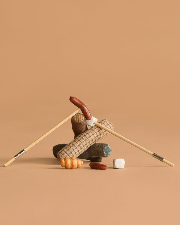 Two chopsticks form an upside-down V over log-shaped fabric, a plush croissant, and marshmallows—this whimsical Maileg Bonfire set inspires imaginative camping adventures, displayed on a plain beige background.